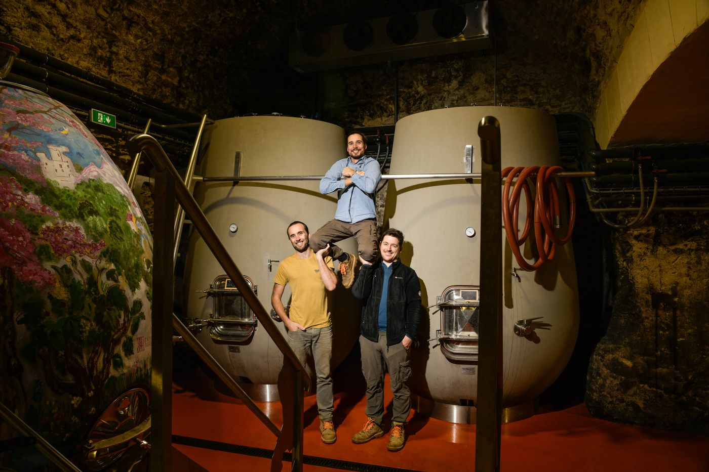 Arthur, Hugo et Jean dans le chai voûté, entre deux grandes cuves en béton, à côté d’une cuve peinte